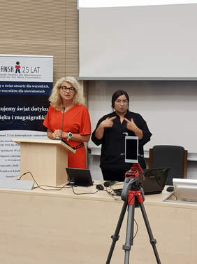 Pokaż zdjęcie: od lewej kobieta w czerwonej sukience stoi przed mikrofonem za mównicą, obo kobieta ubrana na czarno tłumaczy na język migowy