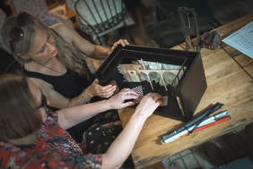 Pokaż zdjęcie: Dwie kobiety siedzą przy stole i dotykają makietę przedstawiającą scenografię na scenie teatru.