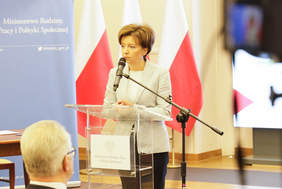 Pokaż zdjęcie: Elegancko ubrana kobieta stojac przy mównicy przemawia do mikrofonu. W tle biało- czerwone flagi narodowe.