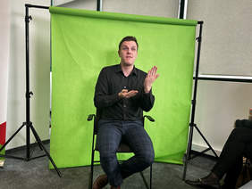 Pokaż zdjęcie: tłumacz języka migowego siedzący na krześle, na zielonym tle