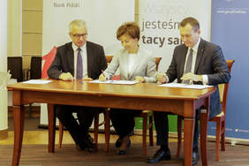 Pokaż zdjęcie: trzy osoby siedzą przy stole. Kobieta siedząca w srodku podpisuje dokument. Męzczyzna po prawej stronie podpisuje drugi egzemplarz dokumentu.
