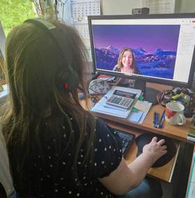 Przy biurku w słuchawkach nausznych siedzi kobieta z długimi włosami, przed nią stoi monitor komputera. Na monitorze widać kobietę.