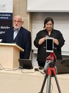 Pokaż zdjęcie: od lewej mężczyzna w garniturze stoi przed mikrofonem za mównicą, obo kobieta ubrana na czarno tłumaczy na język migowy