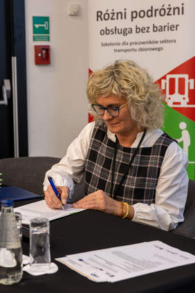 Pokaż zdjęcie: Przedstawicielka Kolei Wielkopolskich p. Joanna Skubiszyńska siedzi przy stole i podpisuje deklarację dotyczącą Partnerstwa na rzecz dostępnej obsługi różnych podróżnych. W tle baner z napisem Różni podróż – obsługa bez barier.