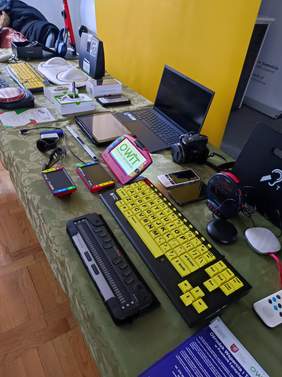 Pokaż zdjęcie: Stół na którym leżą klawiatury, laptopy, czytniki, powiększalniki, ulotki, smartwatch, linijka brajlowska.