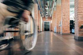 Pokaż zdjęcie: Na zdjęciu: korytarz w budynku, widać fragment osoby na wózku inwalidzkim, wózek jest w ruchu (fot. pexels.com)