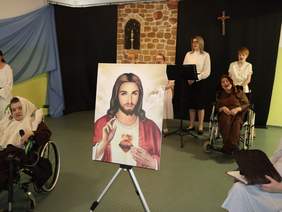 Pokaż zdjęcie: Na środku sali postawiono obraz Jezusa Miłosiernego, obok obrazu siedzi mężczyzna na wózku, z tyłu za obrazem stoją trzy kobiety i śpiewają.
