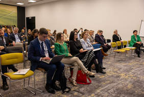 Pokaż zdjęcie: Widok sali konferencyjnej. Kobiety i mężczyźni siedzą na krzesłach ustawionych w rzędach. Jedni patrzą przed siebie, inni czytają dokumenty