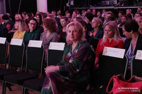 Pokaż zdjęcie: Zdjęcie widowni, na krzesłach siedzą zaproszone osoby, na pierwszym tle kobieta blondynka w zielonej sukience.