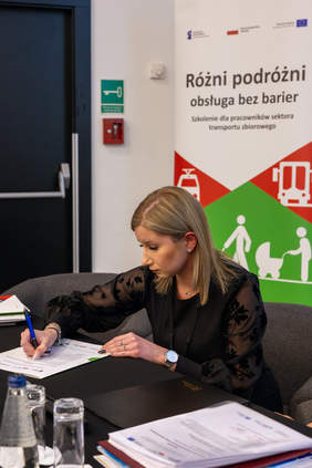 Pokaż zdjęcie: Przedstawicielka Zarządu Transportu Metropolitalnego p. Katarzyna Ziomek siedzi przy stole i podpisuje deklarację dotyczącą Partnerstwa na rzecz dostępnej obsługi różnych podróżnych. W tle baner z napisem Różni podróż – obsługa bez barier.