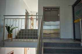 Pokaż zdjęcie: Na holu, nad schodami, winda z blachy z rozsuwanymi drzwiami. Po prawej stronie drzwi szklane wejściowe.