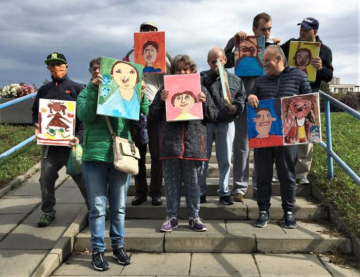 Pokaż zdjęcie: Grupa osób dorosłych niepełnosprawnych, biorących udział w projekcie "Szafirowa Pracownia" Wyspiańskiego. Uczestnicy stoją na schodach, trzymają swoje autoportrety namalowane farbami.