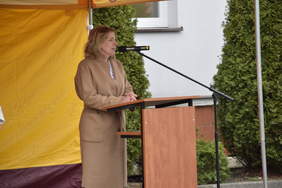 Pokaż zdjęcie: Na dworze, pod żółtym namiotem chroniącym od deszczu stoi mównica. Przy mównicy stoi kobieta blondynka w beżowym płaszczu.