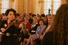Pokaż zdjęcie: Kobieta grająca na skrzypach, sala zamkowa wypełniona gośćmi. 