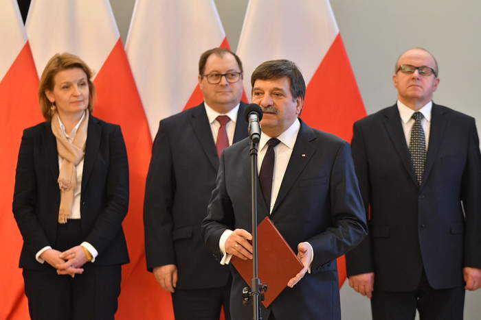 Pokaż zdjęcie: Mężczyzna z teczką w dłoni mówi do mikrofonu, za nim stoją kobieta i dwaj mężczyźni. Tło stanowią biało-czerwone flagi.
