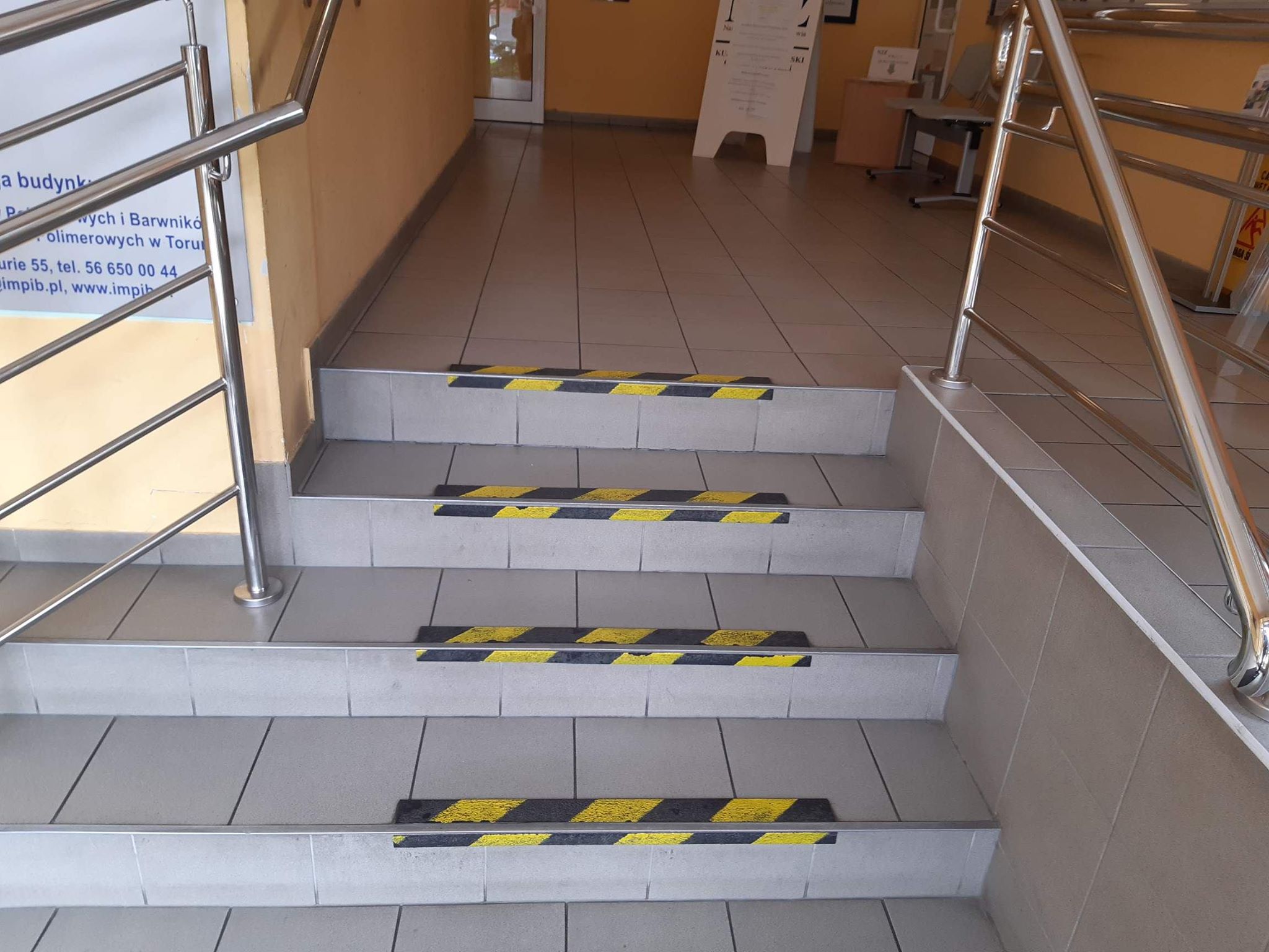 Schody obłożone jasnoszarymi płytkami, na krawędziach schodów czarno-żółte taśmy mające skontrastować stopnie schodów. Po lewej i po prawej stronie widzimy poręcze wykonane ze stali nierdzewnej. Po lewej stronie za poręczą widać tablicę informacyjną. Schody prowadzą na poziom, gdzie widzimy białą tablicę informacyjną i drzwi po lewej stronie.
