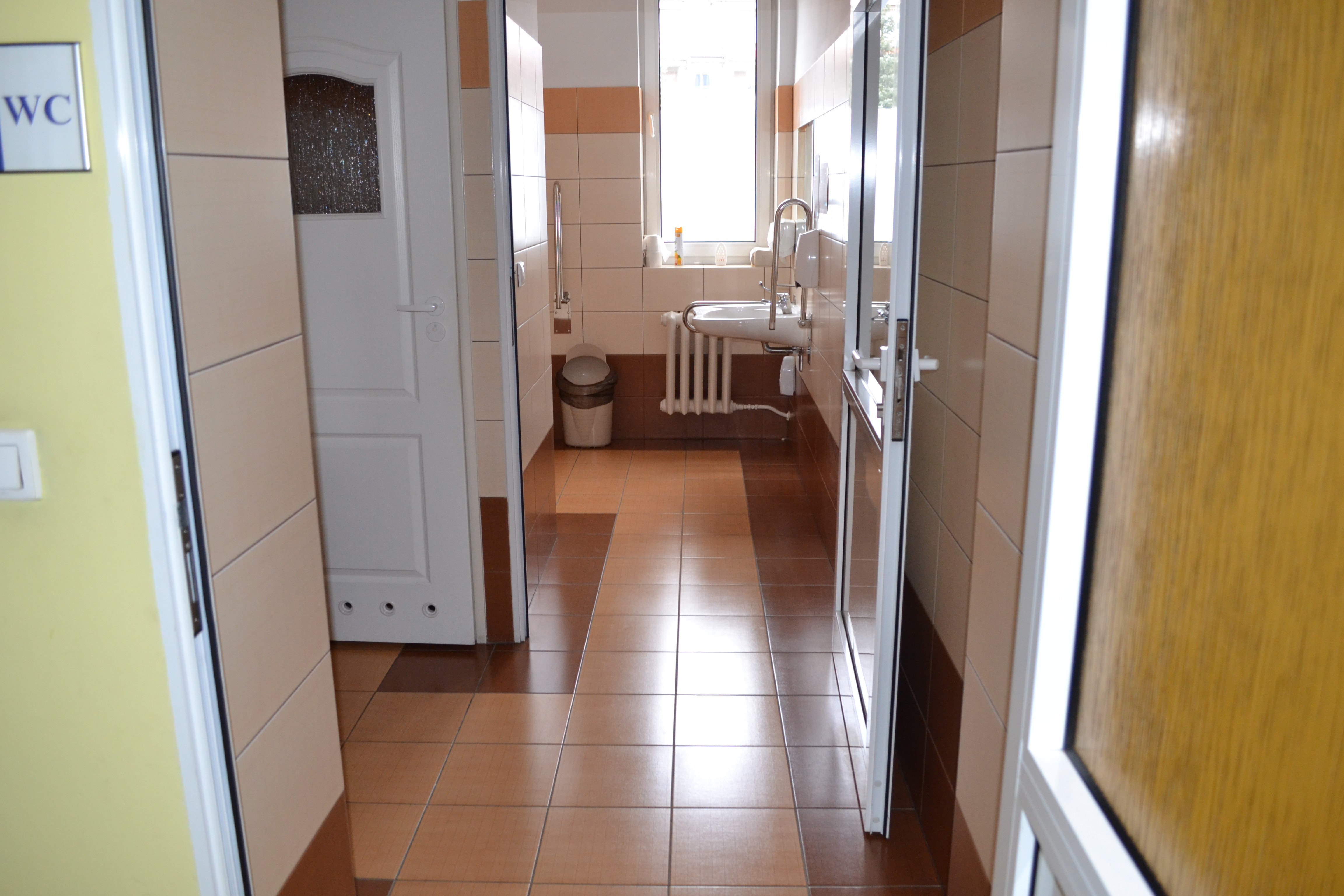 Toaleta z podłogą wyłożona brązowymi płytkami, po lewej stronie widoczne drzwi do kabiny, na wprost widoczne okno i umywalka z uchwytami po obu stronach.