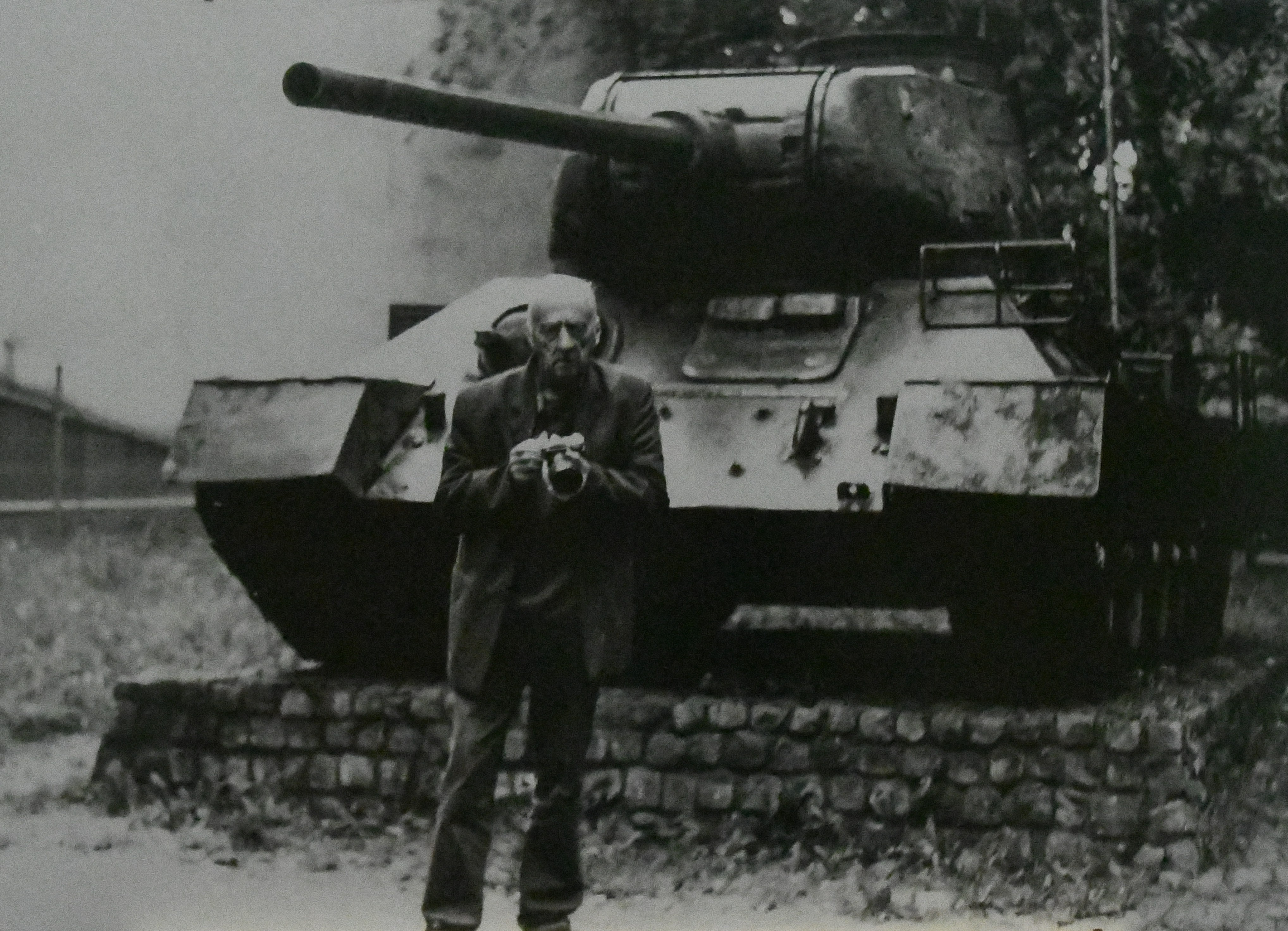 Fotografia czarno biała. Na pierwszym planie starszy mężczyzna z aparatem fotograficznym. W tle czołg z czasów II wojny światowej. Tytuł zdjęcia nawiązuje do potocznego określenia wykonywania zdjęć – strzeliłem fotkę.