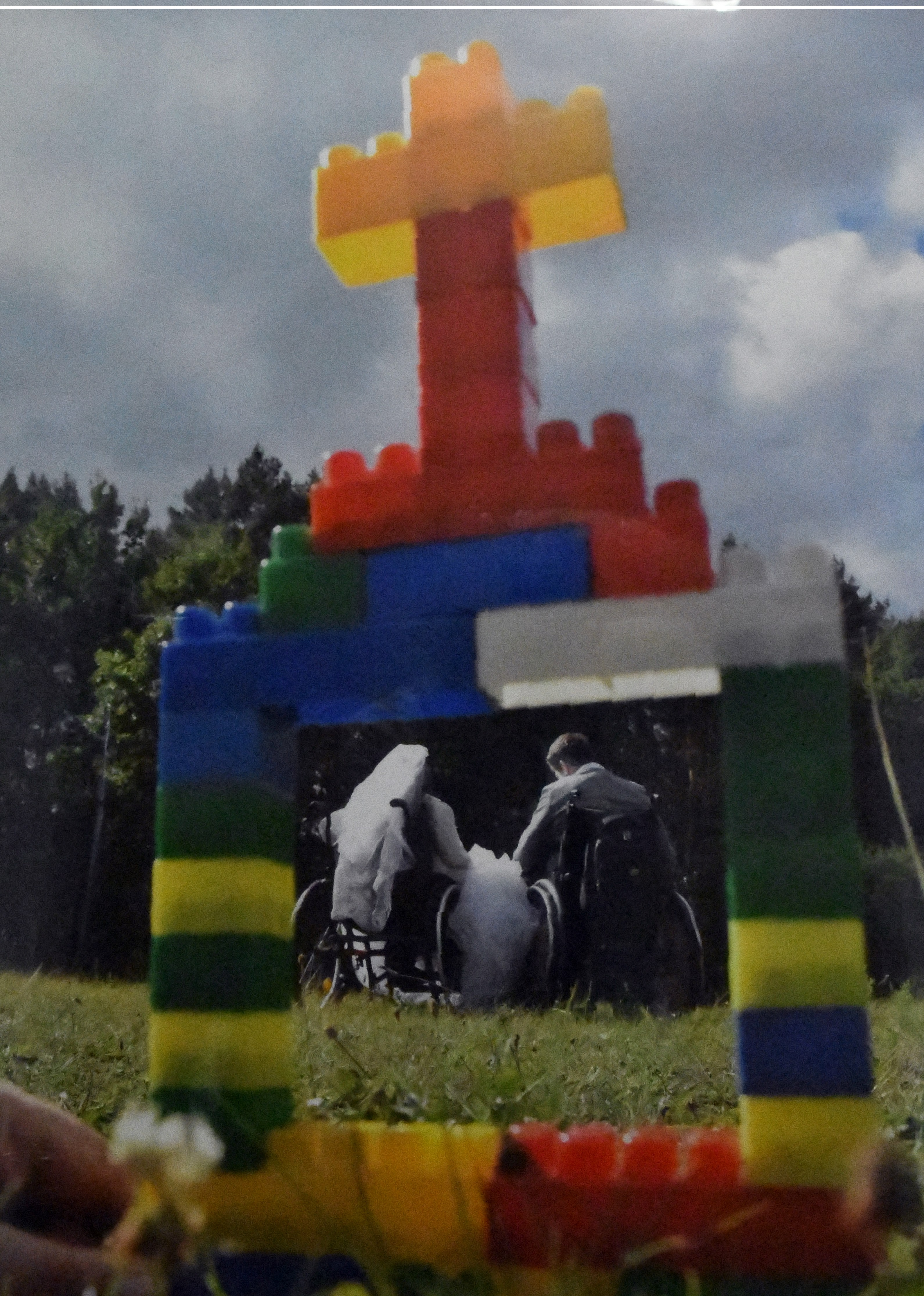 Tło zdjęcia stanowi łąka oraz las w tle. Na pierwszym planie ramka zbudowana z kloców imitująca świątynię. W centrum ramki para młodych na wózkach ubrana w ślubne ubrania.