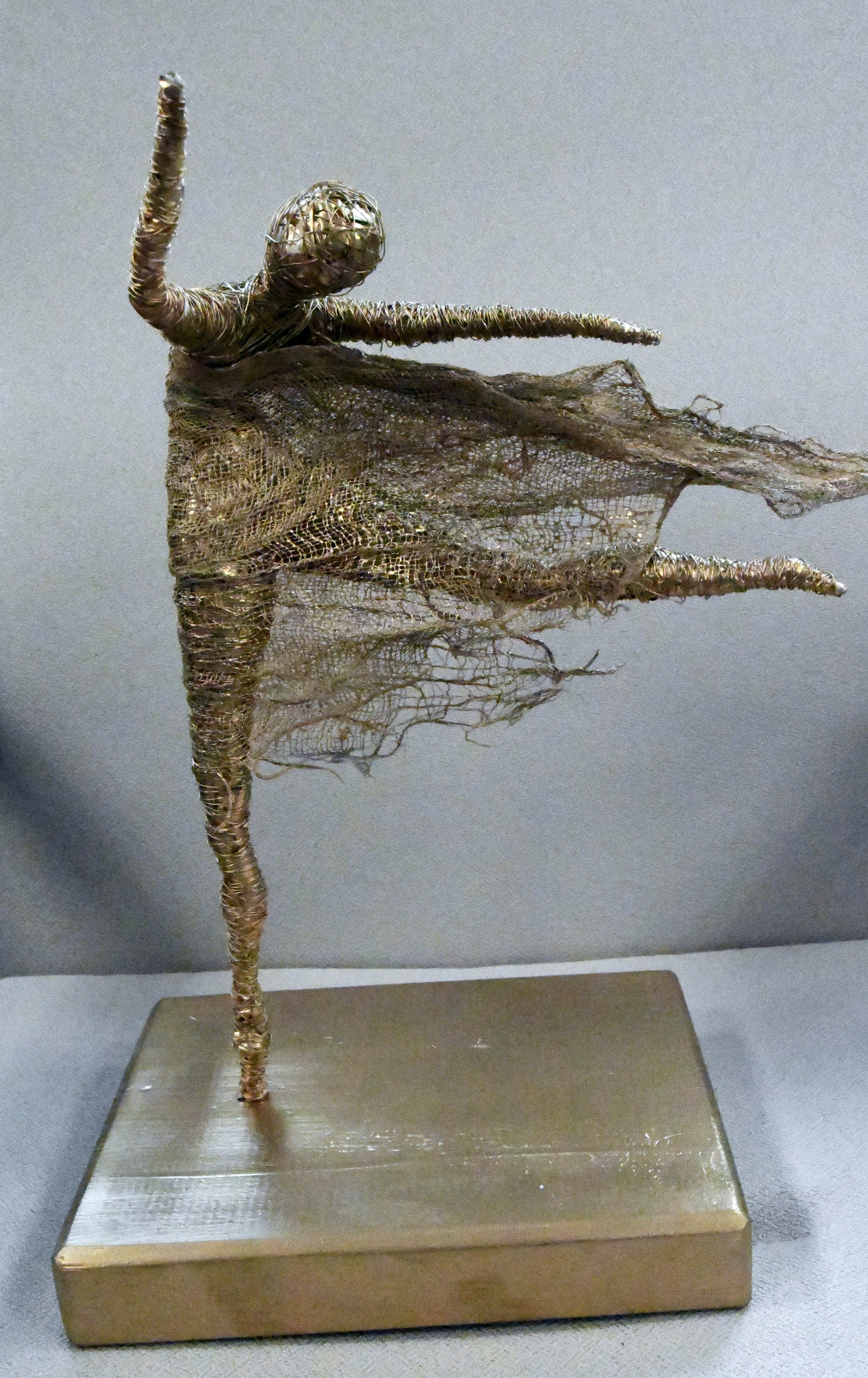 Na kwadratowej podstawie, uproszczona sylwetka baletnicy w ruchu. Postać wykonana metodą, wielokrotnie nawijanych cienkich drucików. Postać stoi na jednej nodze, druga prostopadle do podstawy, ręce uniesione do góry. Struj postaci zwiewany wiatrem od lewej do prawej strony rzeźby.