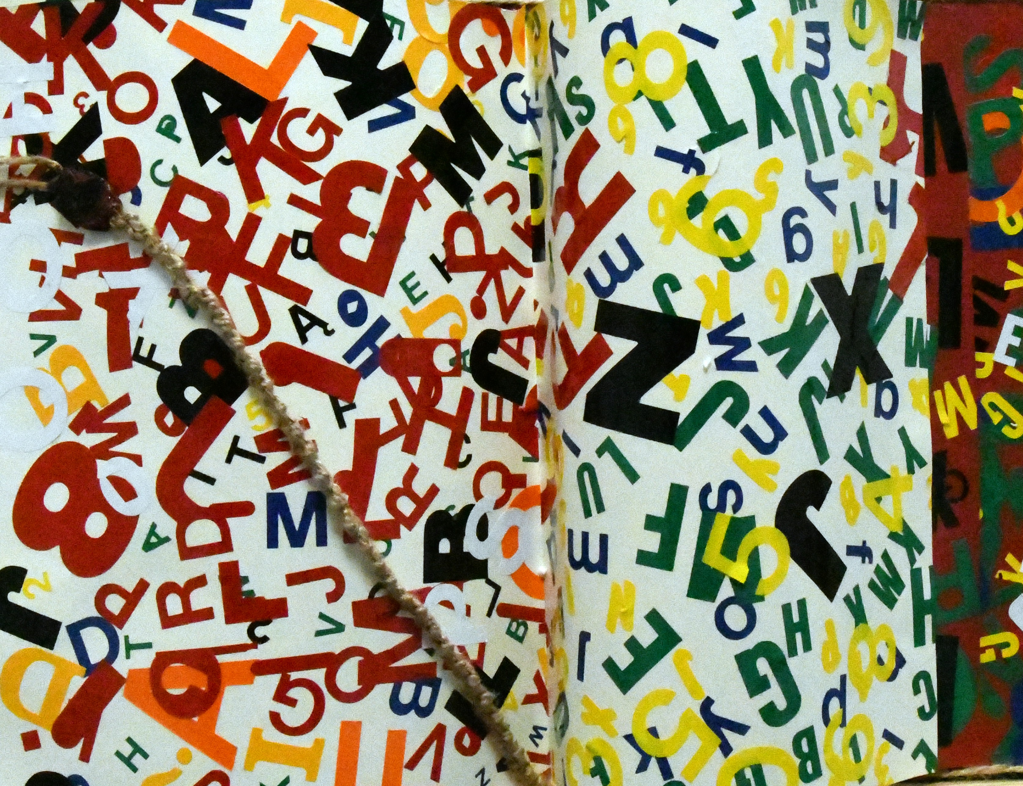 Przestrzenna forma z papieru, przedstawiająca uniesione strony książki. Na stronach książki umieszczone różnokolorowe litery o różnej wielkości. Strony przepasane sznurem, który jest zakończony pieczęcią lakową.