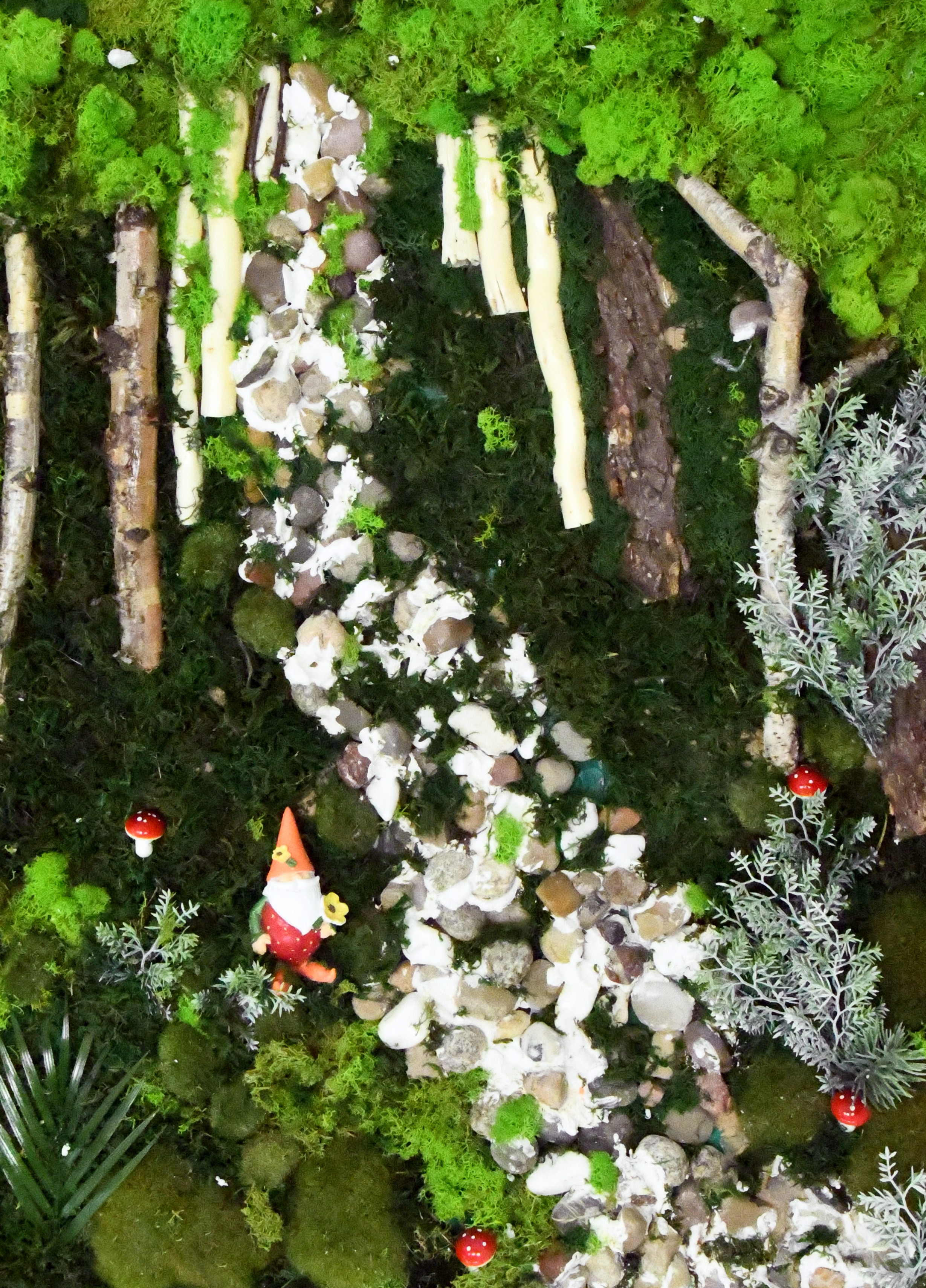 Na płaskiej prostokątnej podstawie umieszczone różnorodne materiały: kamyczki, tkanina, kora z drzewa, gotowe plastikowe elementy . Całość przedstawia strumień, obok którego stoi krasnal wśród muchomorów. Strumień przepływa przez las.