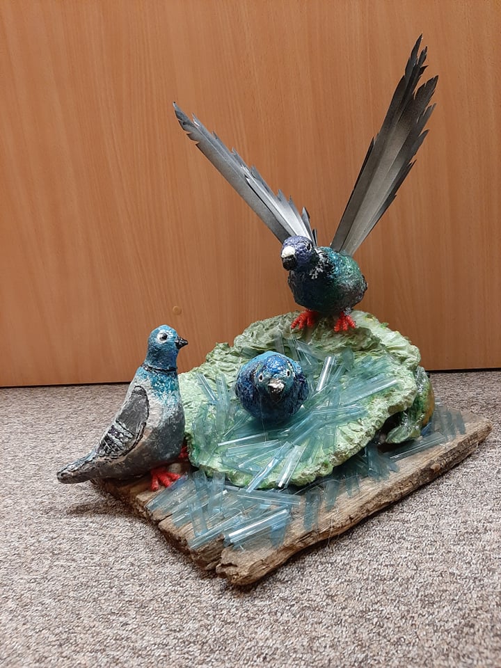 Rzeźba przedstawia ptasie gniazdo. W środku gniazda siedzi pisklę. Na górnej krawędzi siedzi ptak z uniesionymi skrzydłami, przy dolnej krawędzi siedzi drugi ptak, ale skrzydła ma złożone