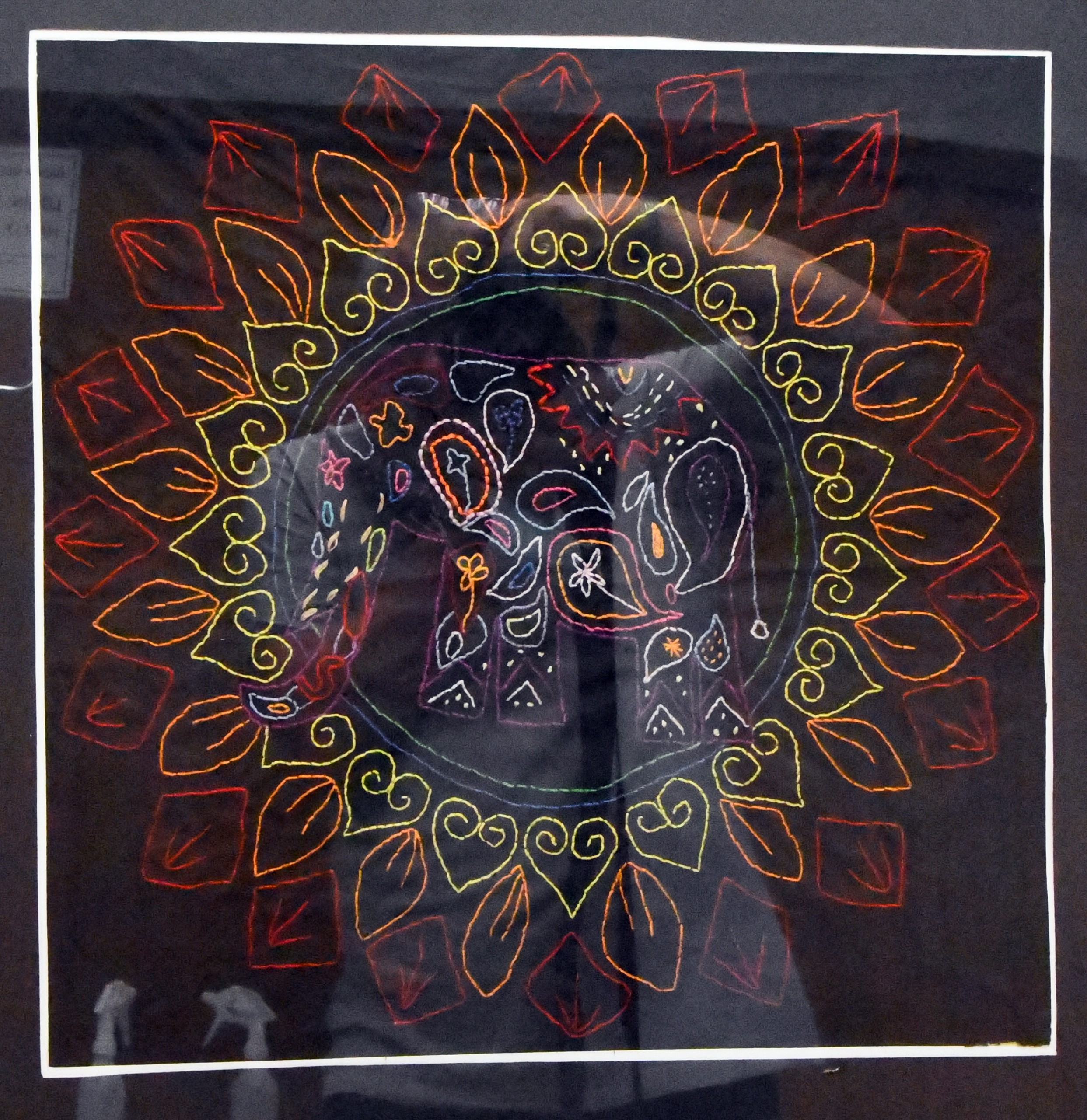 Wyszywane koło. W środku wielokolorowe figury. Koło otaczają trzy pierścienie figur. Każdy z pierścieni jest w innym kolorze i przedstawia różny znak graficzny.