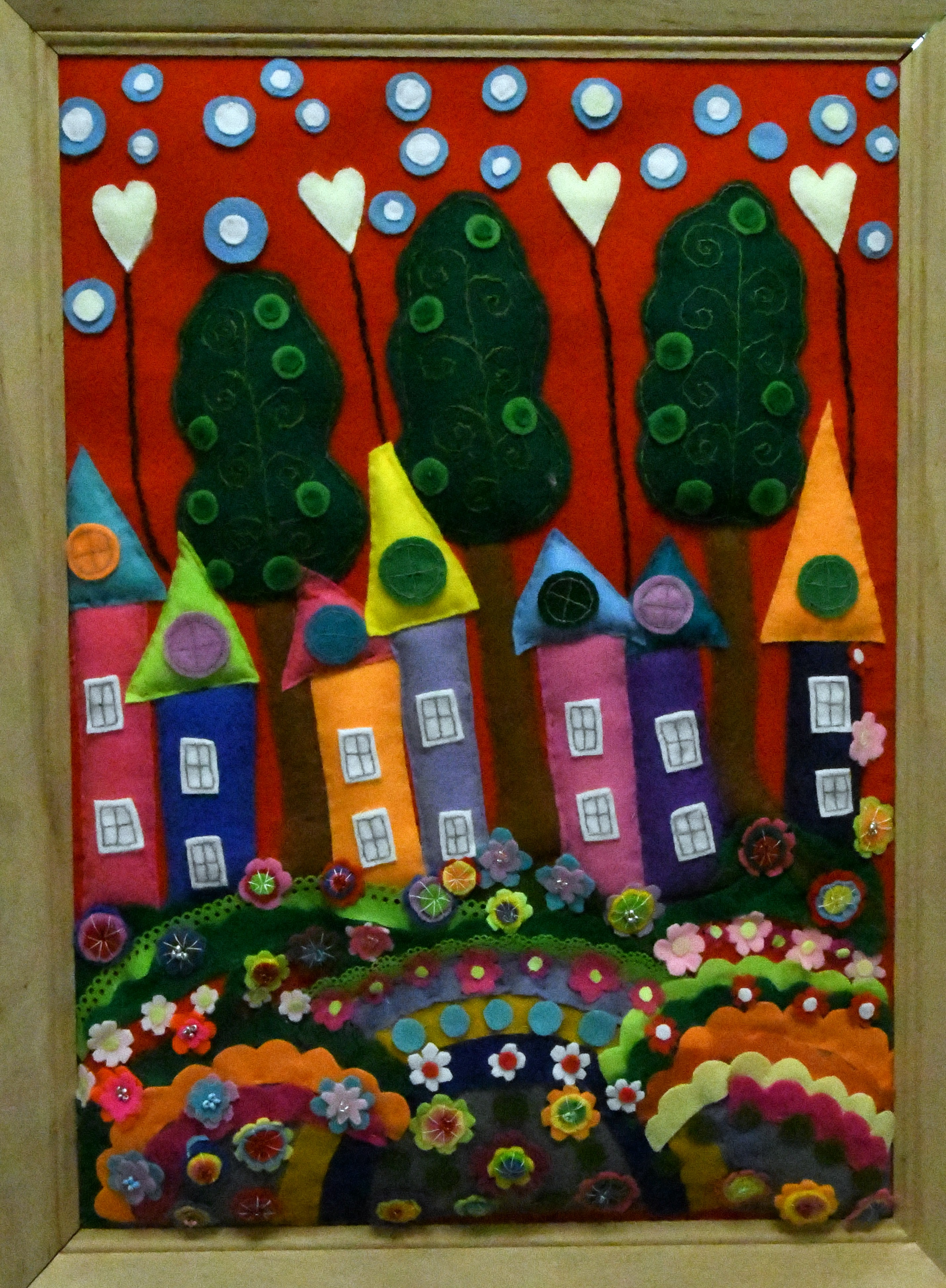 Wielokolorowa, wypukła tkanina. Siedem wielokolorowych piętrowych domków na tle drzew. Domki ustawione nieregularnie na różnej wysokości wzniesień. Na wzniesieniach różnokolorowe kwiaty. Niebo w kolorze czerwonym z chmurami w postaci niebieskich okręgów.