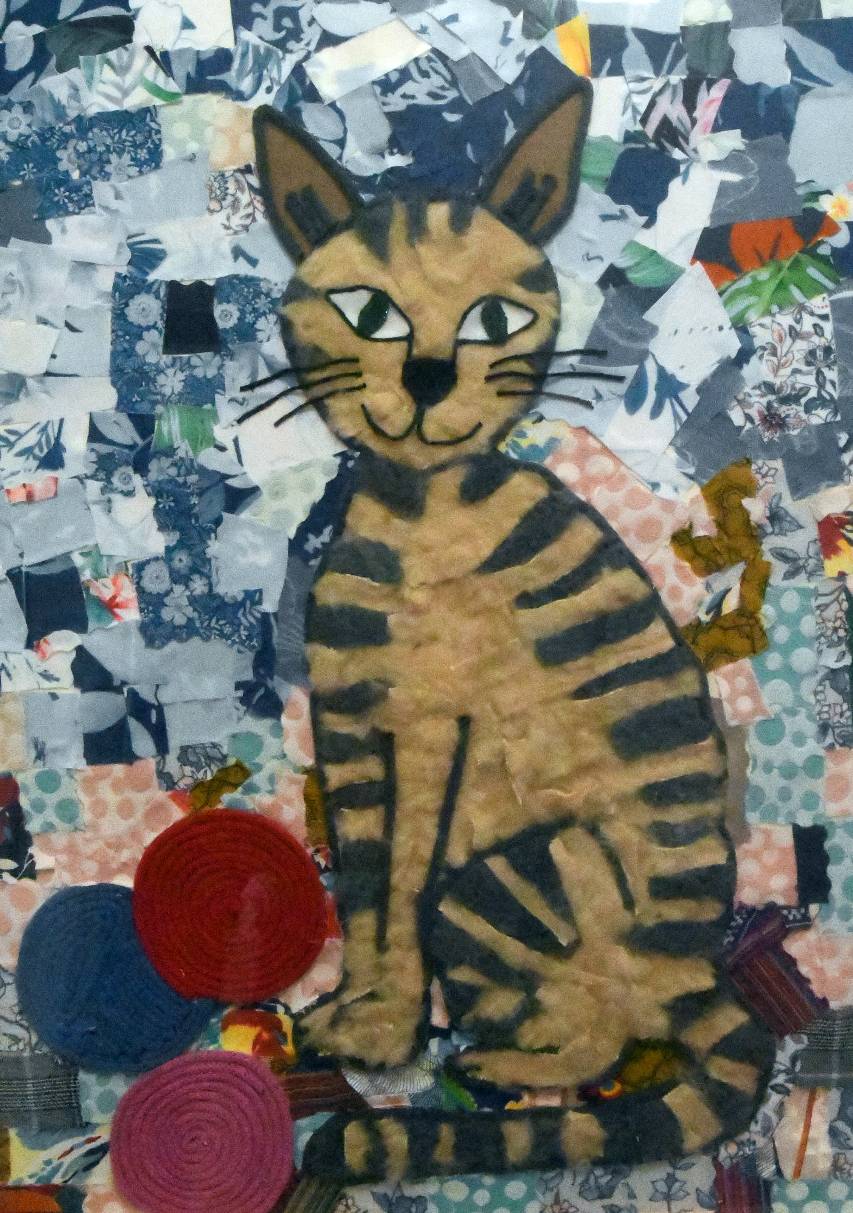 Postać kota wyklejona z różnych tkanin o różniej grubości, dzięki czemu uzyskano wypukłości. Tło jest wielokolorowe, a pomiędzy łapami kota trzy kłębki włóczki. 