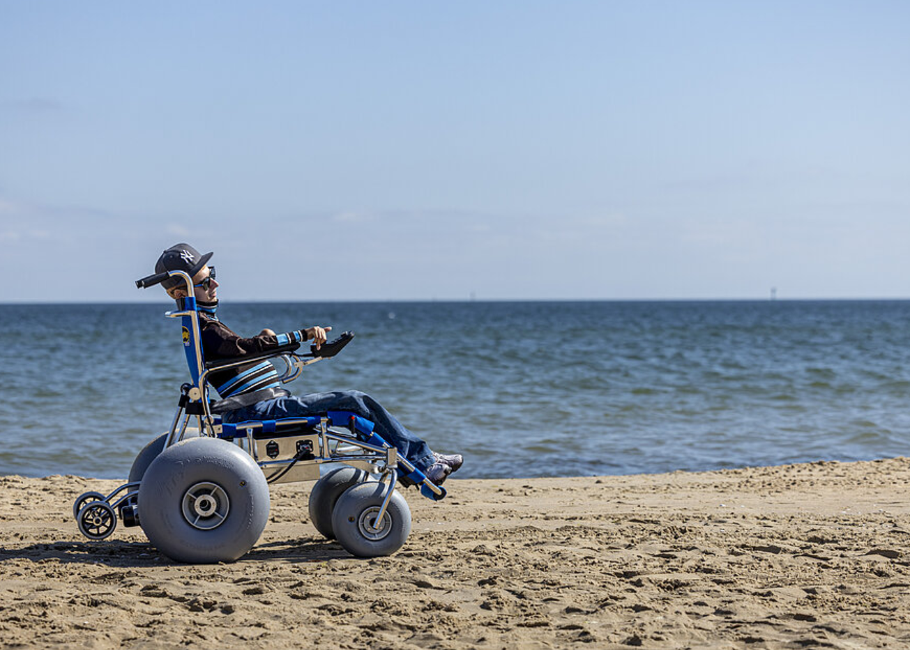 Osoba siedzi na specjalistycznym wózku plażowym z dużymi, szerokimi kołami na piaszczystej plaży. W tle widoczne jest morze i jasne niebo. Wózek ustawiony jest bokiem.