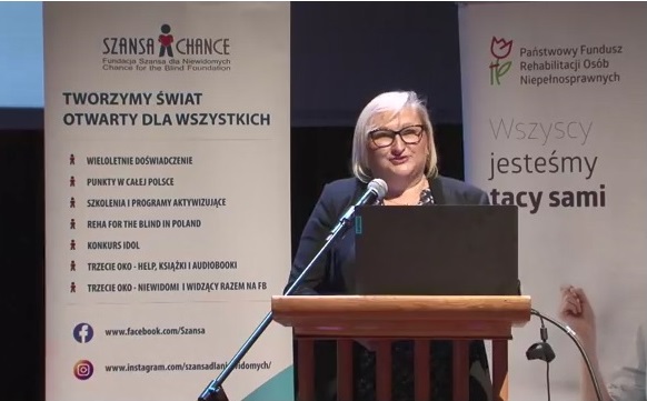 Pokaż zdjęcie: Kobieta z włosami koloru blond, nosząca okulary i ubrana w czarną marynarkę mówi do mikrofonu z tzw. ambony, na której stoi laptop. W tle widoczne są banery Fundacji Szansa dla Niewidomych oraz Państwowego Funduszu Rehabilitacji Osób Niepełnosprawnych.