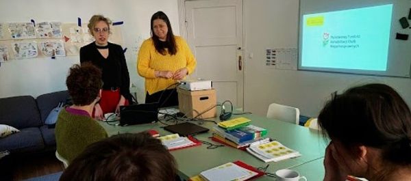 Pokaż zdjęcie: Dwie kobiety stoją za stolikiem, na którym wyłożone są materiały informacyjne. Przy stole siedzą także trzy inne osoby, które przysłuchują się wystąpieniu. Po prawej stronie na ekranie wyświetlony jest duży logotyp PFRON.