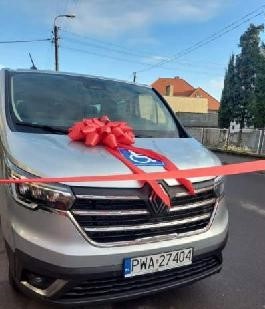 Pokaż zdjęcie: Srebrny bus z czerwoną kokardą na masce pojazdu. W tle dom jednorodzinny, drzewa iglaste, jezdnia, lampa uliczna oraz prawie bezchmurne niebo.
