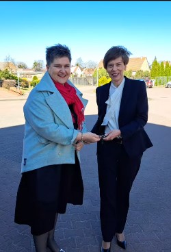 Kobieta w czarnym garniturze wręcza klucze do nowego busa kobiecie w jasnoniebieskim płaszczu. Spotkanie odbywa się na zewnątrz, w tle widać zabudowania i zieleń.