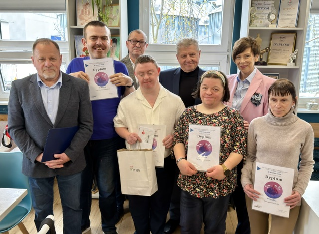 Pokaż zdjęcie: Zdjęcie grupowe ośmiu osób, z których część trzyma pamiątkowe dyplomy laureatów i uczestników XXII edycji wojewódzkiego etapu Konkursu Plastycznego „Sztuka Osób Niepełnosprawnych”.