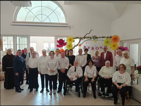 Pokaż zdjęcie: Grupa osób w eleganckich strojach pozuje w dwóch rzędach w jasnym pomieszczeniu. W tle dekoracje z dużych papierowych kwiatów i napis „PFRON”. Jeden z mężczyzn ma na sobie charakterystyczną czerwoną marynarkę.