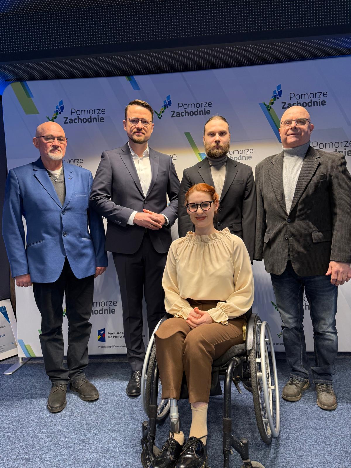 Portretowe zdjęcie pięciu osób pozujących na tle ścianki z logo „Pomorze Zachodnie”. W centrum siedzi uśmiechnięta kobieta w okularach na wózku inwalidzkim, ubrana w beżową bluzkę z falbaną przy szyi i brązowe spodnie. Za nią stoi czterech mężczyzn w marynarkach. Całość kompozycji jest jasna i oficjalna. Na dole ścianki widoczne są logotypy Funduszy Europejskich.