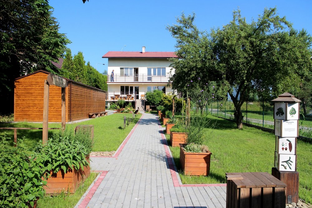 Widok chodnika z kostki brukowej prowadzącego do budynku. Wzdłuż niego gazony z roślinami, drewniana altana i drzewa. 