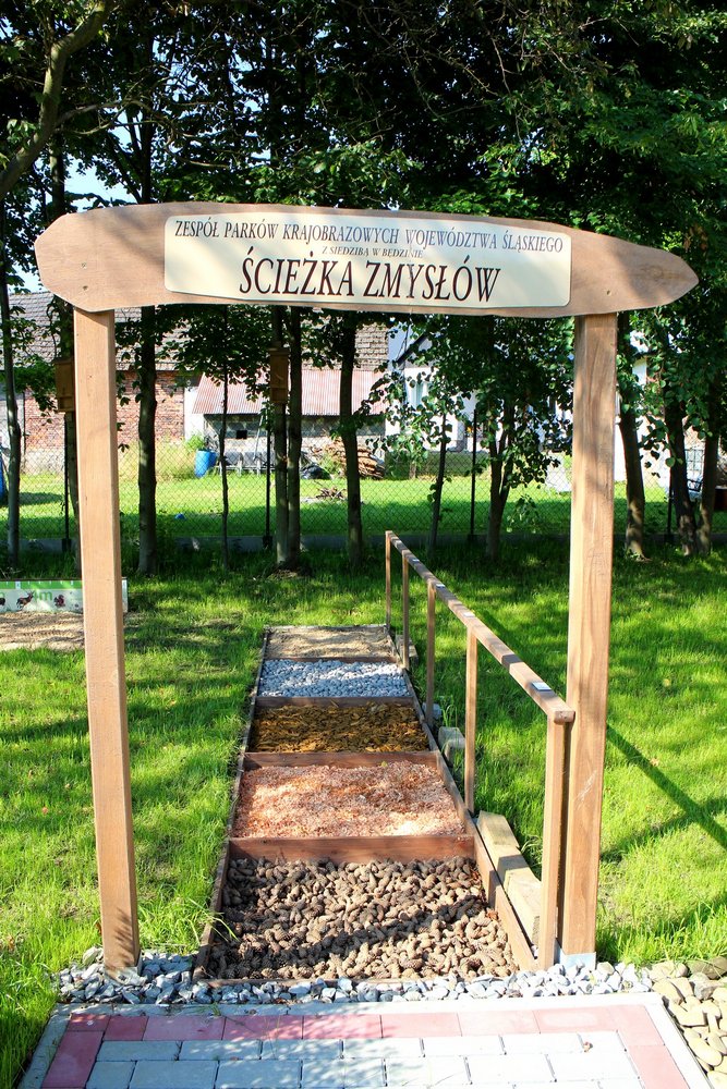 Drewniana tablica z napisem Zespół Parków Krajobrazowych Województwa Śląskiego z siedzibą w Będzinie Ścieżka Zmysłów. Za nią sensoryczna ścieżka z różnymi rodzajami powierzchni np. żwir, piasek, drewno umieszczonymi w kwadratowych boksach. Obok drewniana barierka. 