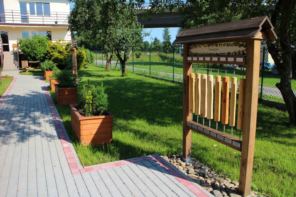 Fragment brukowanego chodnika prowadzącego do budynku, z prawej strony gazony z roślinami oraz tablica edukacyjna Dendrofon.