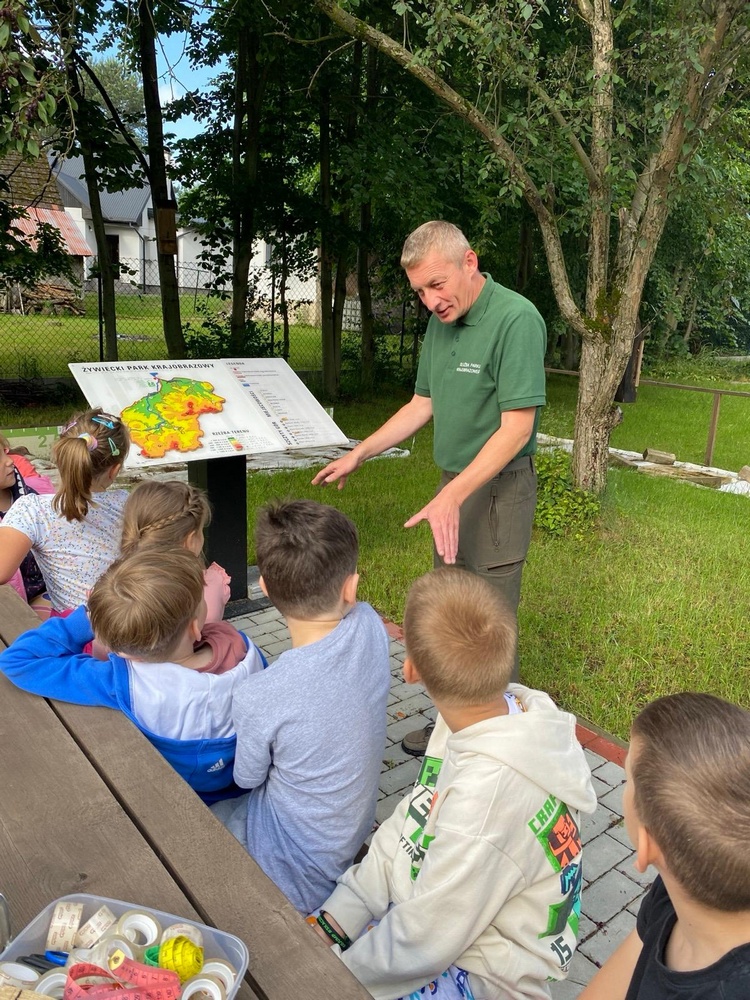 Mężczyzna w stroju pracownika parku krajobrazowego prowadzi zajęcia na świeżym powietrzu grupie dzieci siedzącej na ławce. W tle hipsometryczna mapa Żywieckiego Parku Krajobrazowego na podwyższeniu.