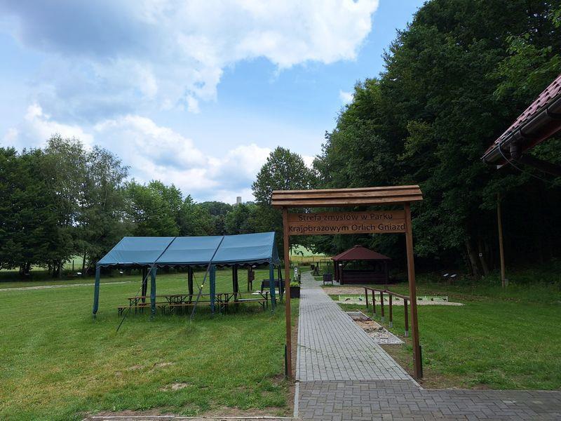 Fragment ścieżki sensorycznej „Strefa zmysłów w Parku Krajobrazowym Orlich Gniazd”. Drewniana tablica z napisem Strefa zmysłów w Parku Krajobrazowym Orlich Gniazd. Za nią chodnik z kostki brukowej. Po lewej zielony namiot z ławkami i stolikami. W tle drzewa.