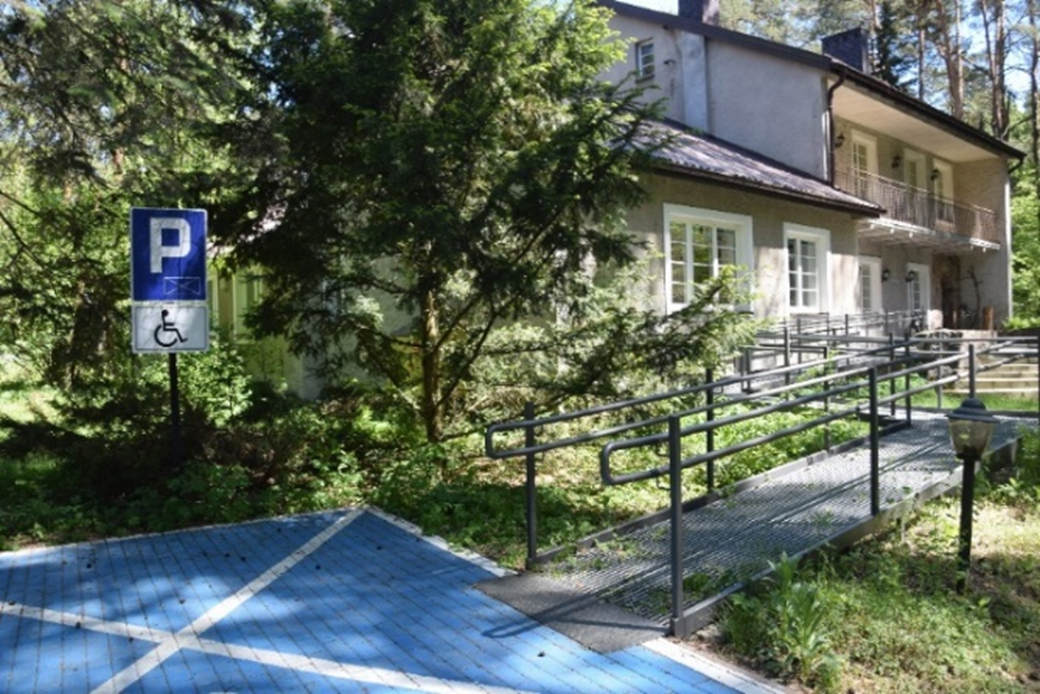 Miejsce parkingowe dla osób z niepełnosprawnościami, z którego do budynku w tle prowadzi metalowy podjazd. Wokół drzewa.