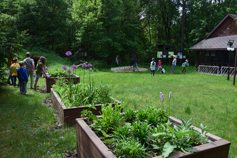 Widok miejsca wypoczynku. Na pierwszym planie podwyższone drewniane skrzynie z różnymi rodzajami roślin. W tle na polanie grupa osób obok drewnianego budynku. Wokół las.