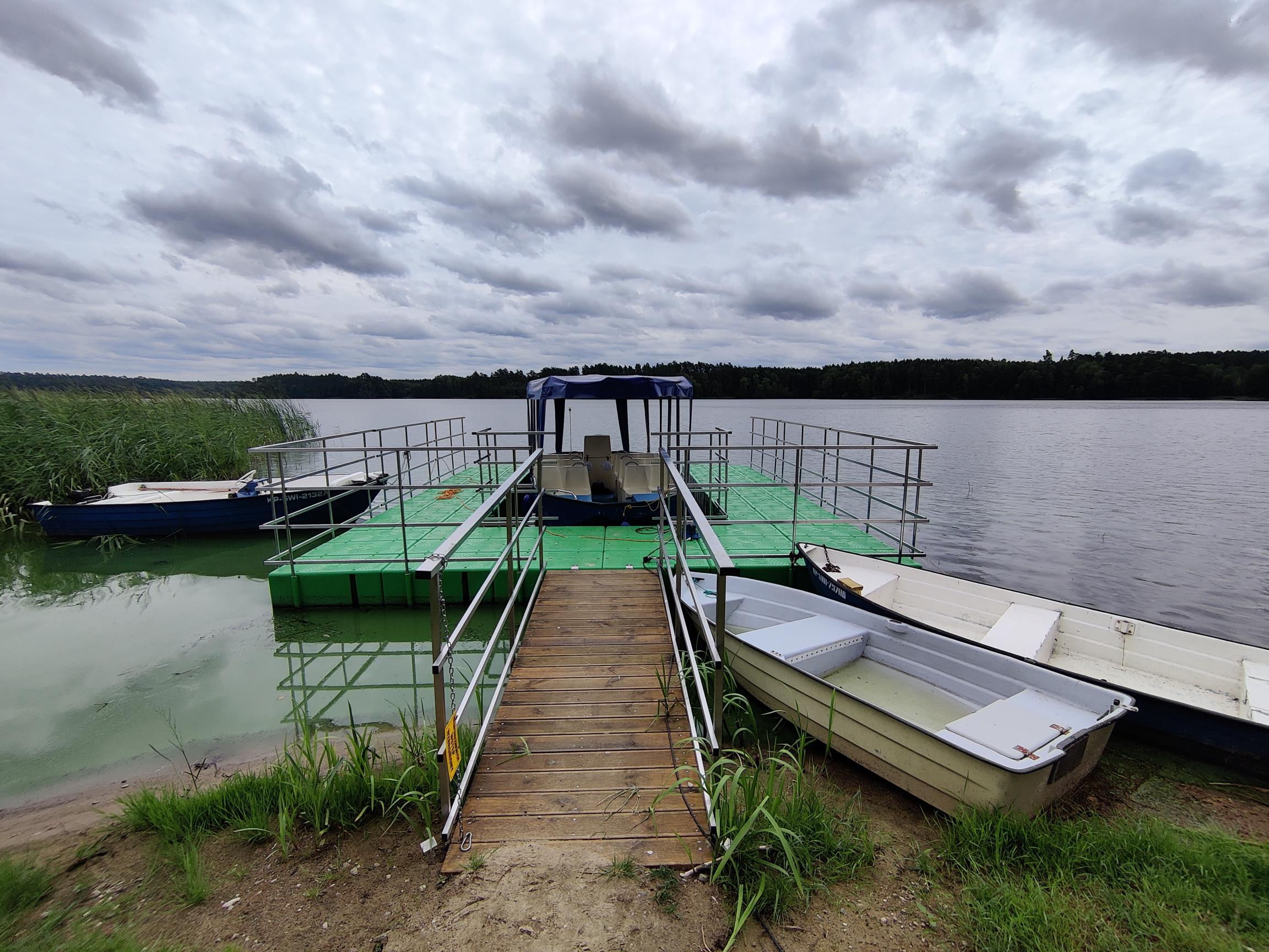 Pomost pływający. Pomost pływający na jeziorze połączony z brzegiem drewnianym podjazdem. Pomost w kształcie litery U zabezpieczony metalowymi barierkami. W środku tramwaj wodny.