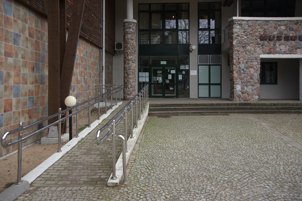 Po lewej stronie podjazd do budynku z kostki brukowej. Na wprost wejście po schodach z przeszklonymi drzwiami. Fasada budynku Muzeum Przyrodniczo-Leśnego BPN zrobiona z kamieni.