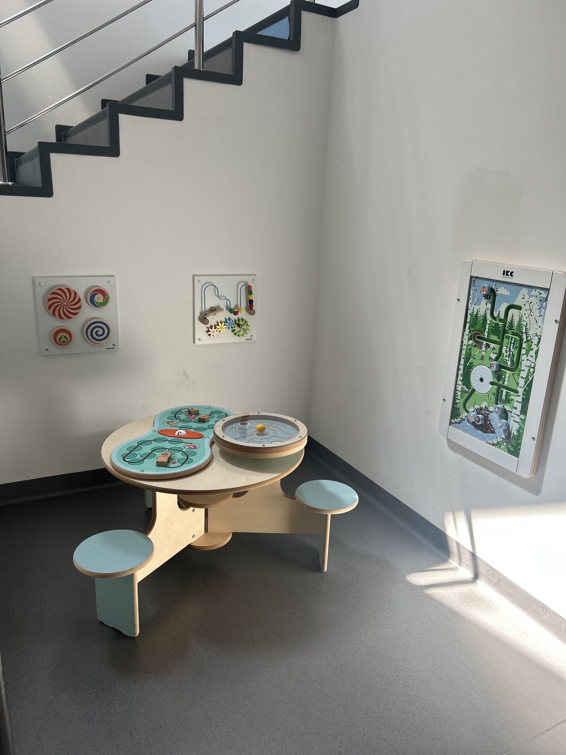 Kącik malucha w Ośrodku Edukacji Przyrodniczej „Młynarzówka”. Kącik malucha w rogu obok schodów. Na środku drewniany stolik interaktywny z siedzeniami. Na ścianach trzy drewniane tablice do zabawy.