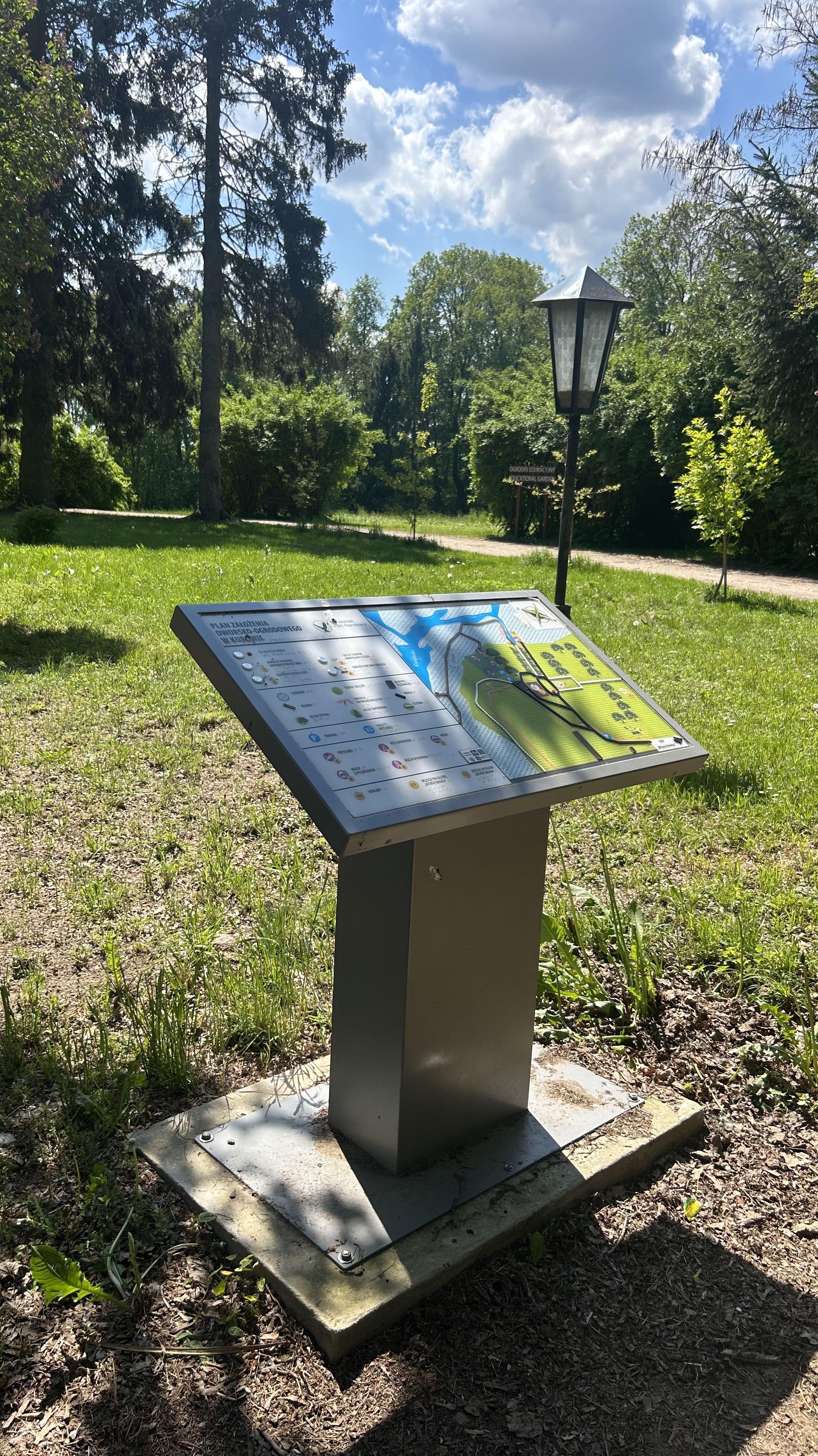Plan tyflograficzny na parku przydworskim w Narwiańskim Parku Narodowym. Plan tyflograficzny parku przydworskiego w Narwiańskim PN na metalowym postumencie. W tle latarnia, ścieżka leśna, trawa, drzewa i błękitne niebo.