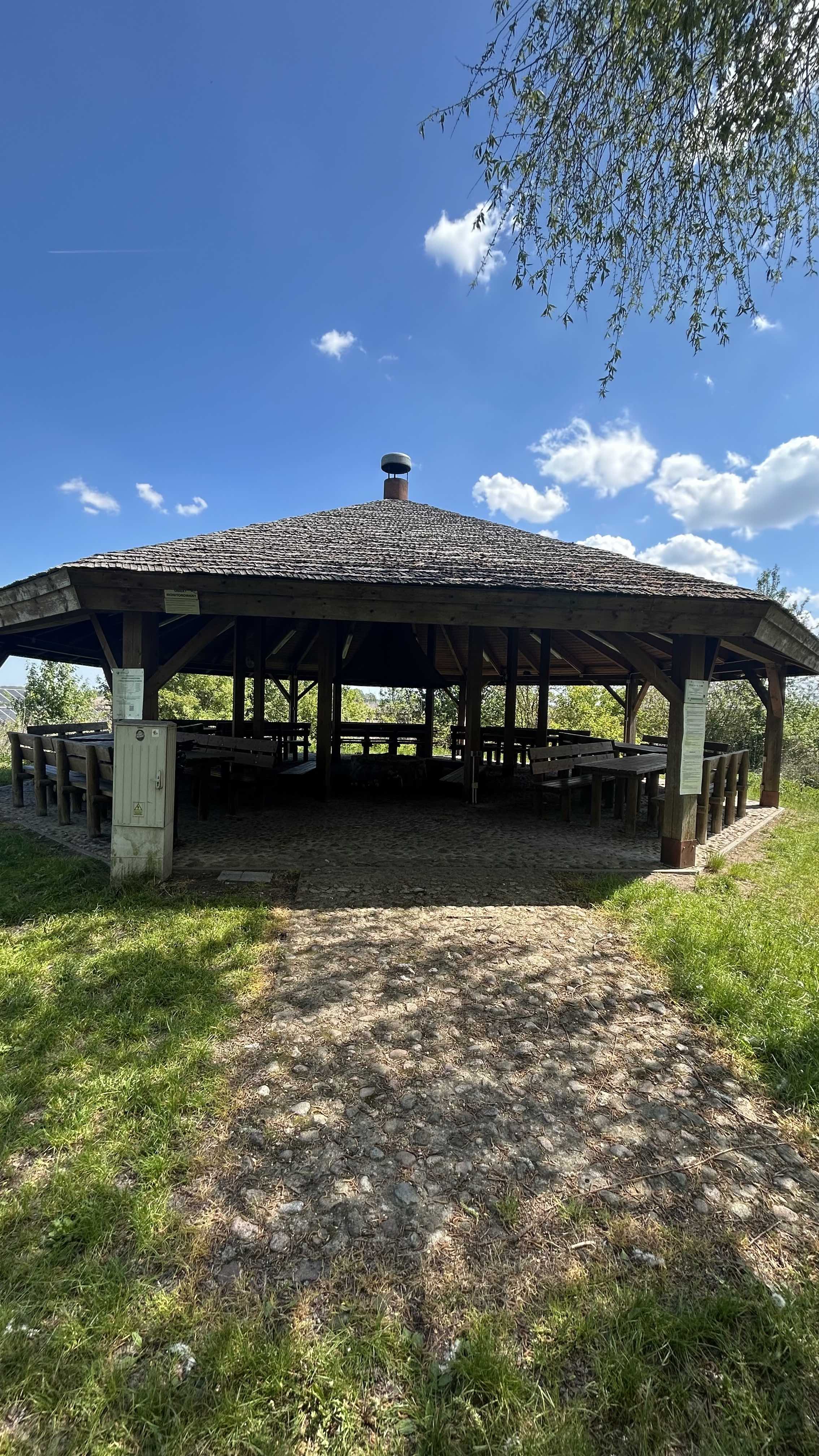 Altana z miejscem na ognisko w Narwiańskim Parku Narodowym. Widok altany z miejscem na ognisko w Narwiańkim PN. Duża drewniana altana pokryta strzechą z utwardzonym podjazdem dla wózków. W tle drzewa i błękitne niebo.